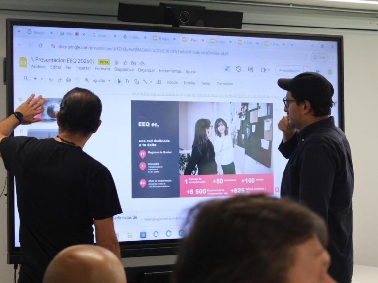 Dos hombres están de pie frente a una pantalla grande. En la pantalla se proyecta una presentación de Google Slides titulada “Presentación EEQ 2026-02”. Uno de los hombres señala la pantalla con la mano mientras el otro observa pensativo con la mano en la barbilla.